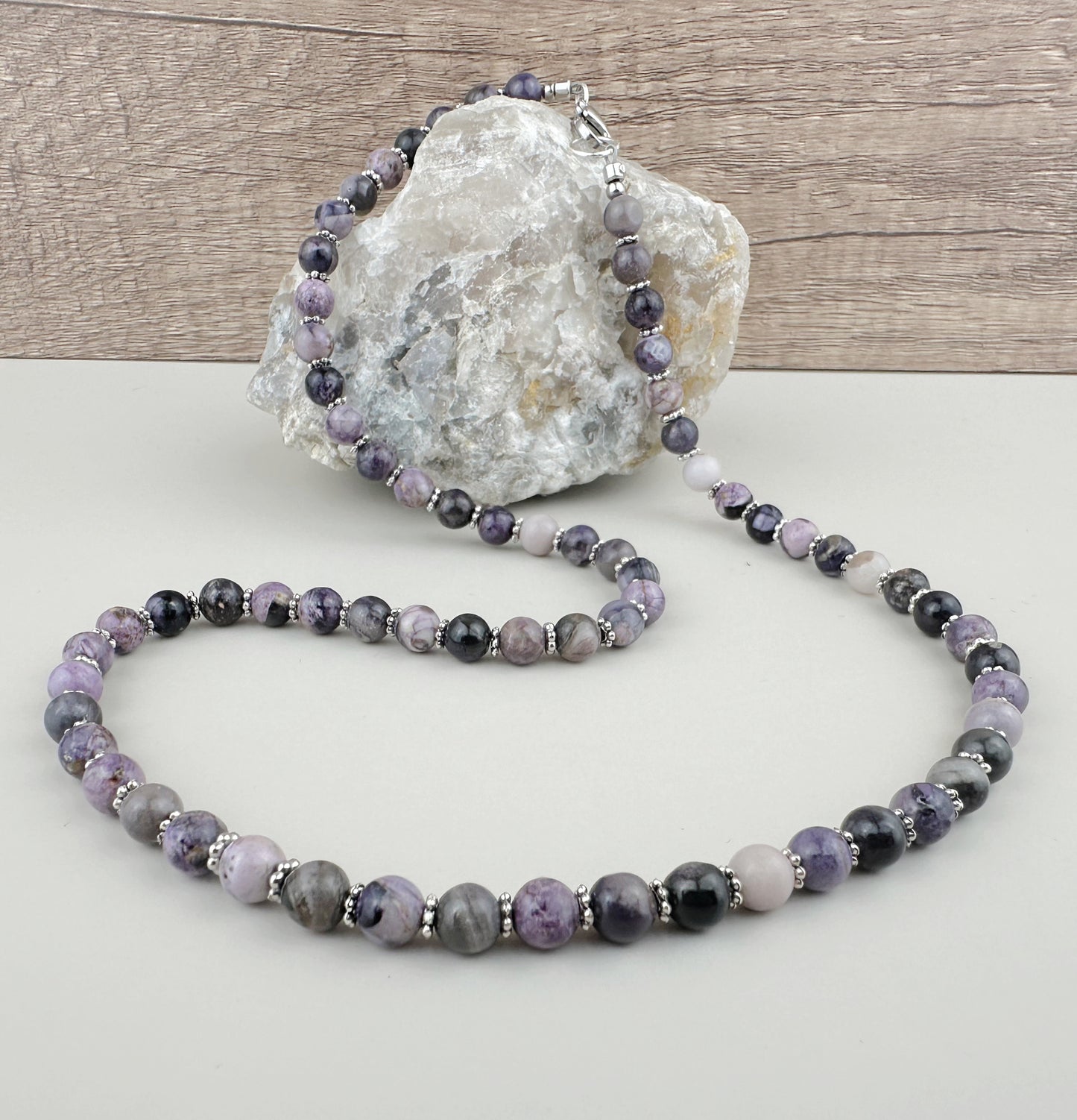 Beaded necklace with purple and gray stones on a white surface with a rock in the background