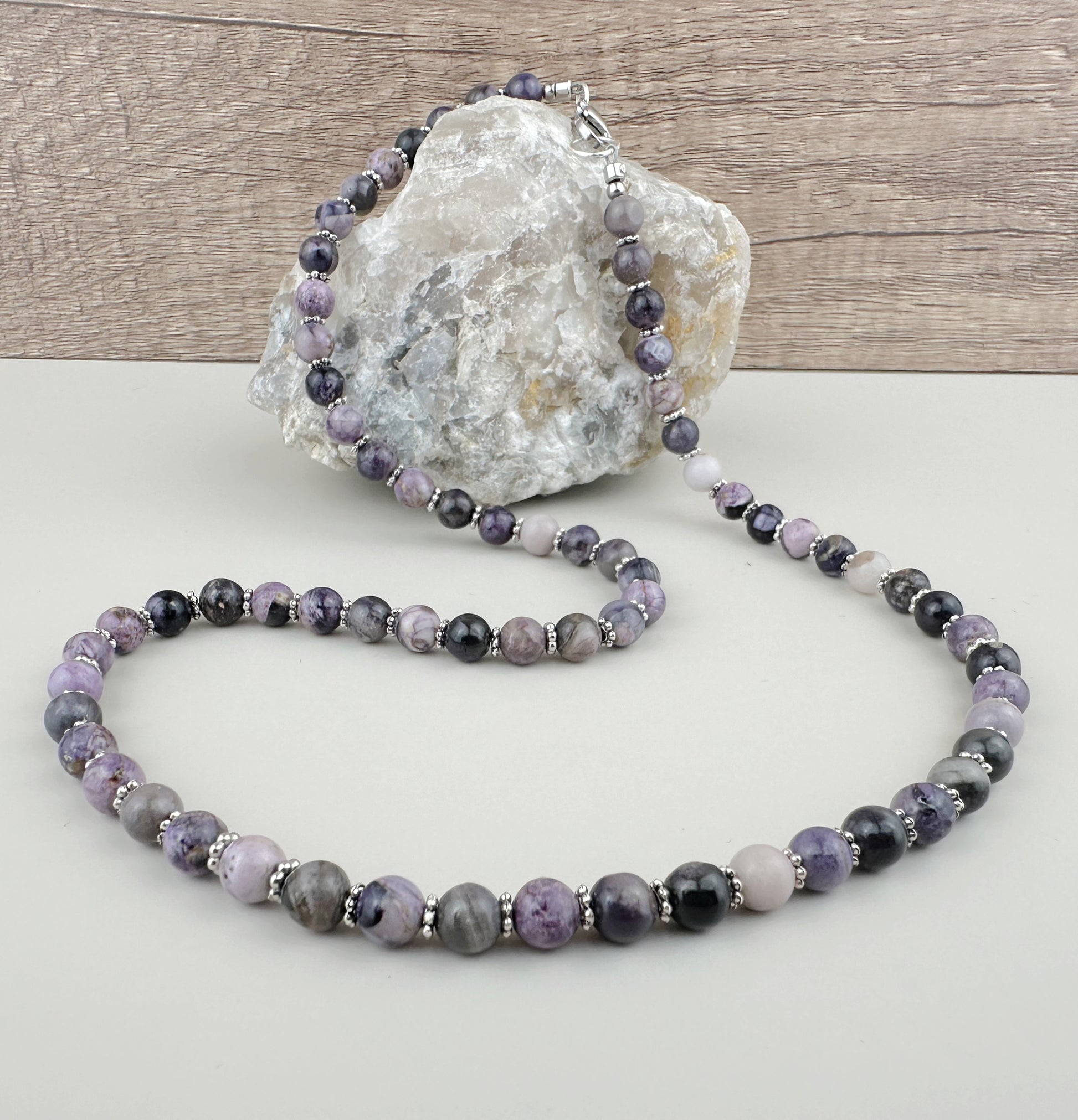 Beaded necklace with purple and gray stones on a white surface with a rock in the background