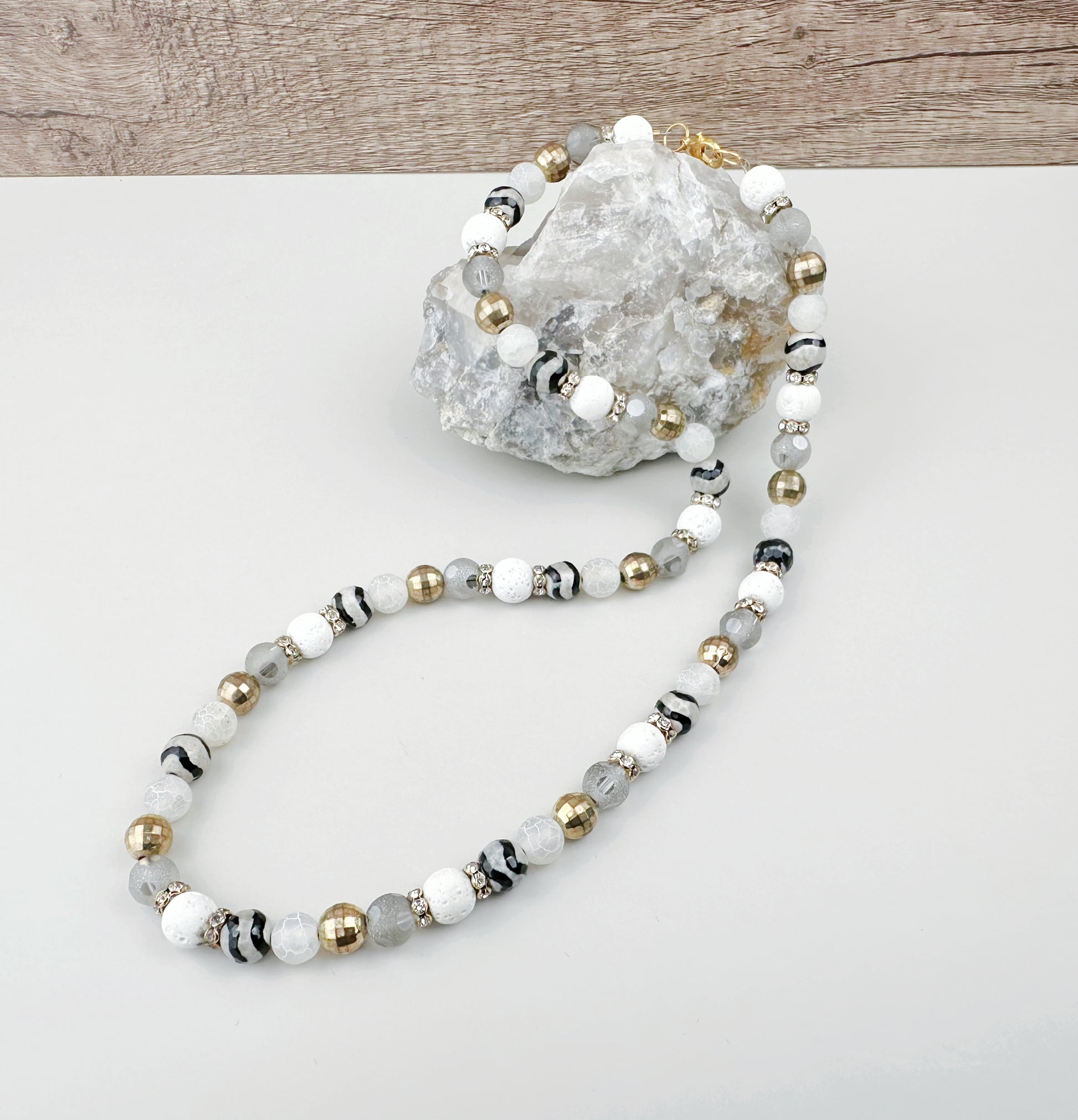 Beaded necklace with white, gold, and gray beads on a white surface with a decorative rock.