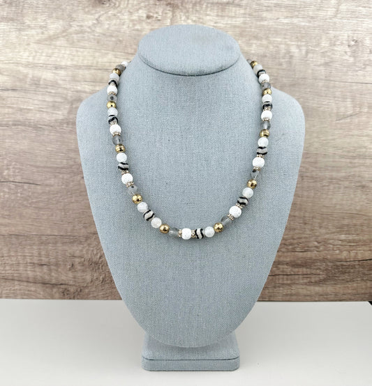 Necklace with white, black, and gold beads on a gray bust against a wooden background